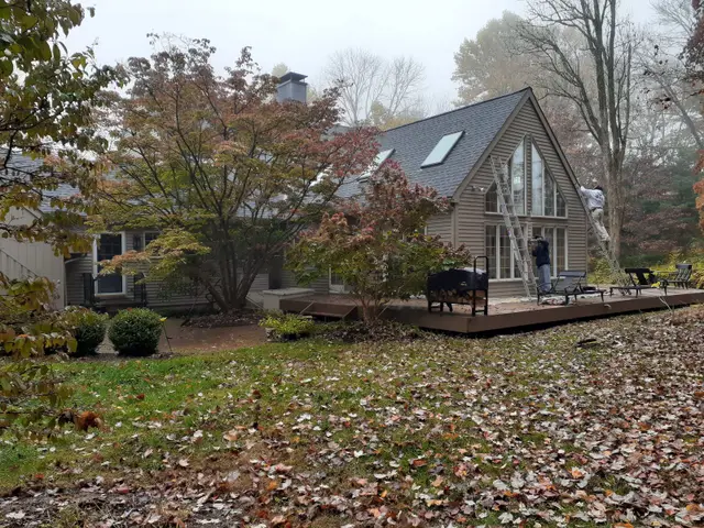 A cedar-sided A-frame house with a wooden deck in autumn leaves, a painter working from a ladder at the gable.