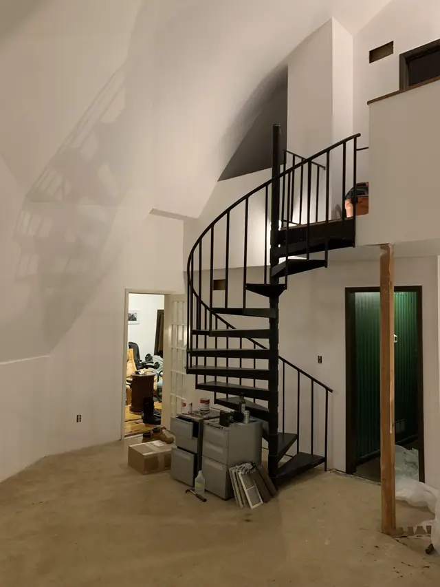 Inside a geodesic dome: angled white walls, a black spiral stair and a green door, the floor still bare.