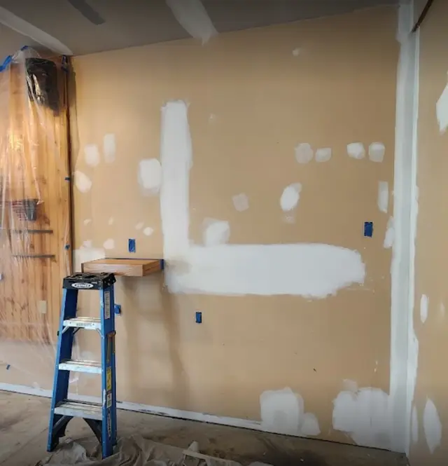A beige wall with a long L-shaped patch and many smaller patches in white compound, a stepladder beside it.