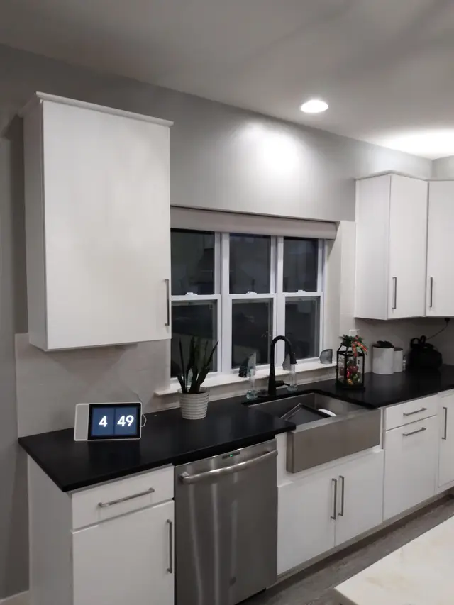 A kitchen with white shaker cabinets, black counters and a farmhouse sink under a window.