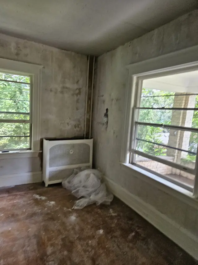 A room in an older house with its walls patched and part-way through a skim coat, a radiator cover under the window.