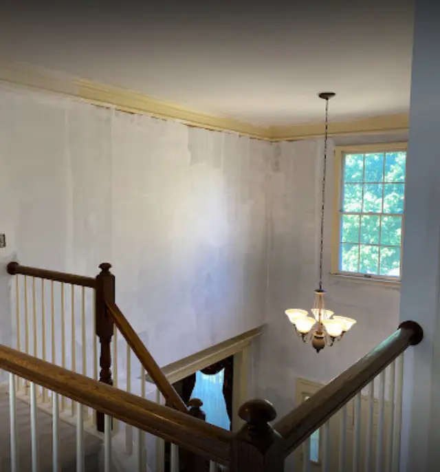 A two-story stairwell wall patched and primed in white over a wood banister, a chandelier hanging below.
