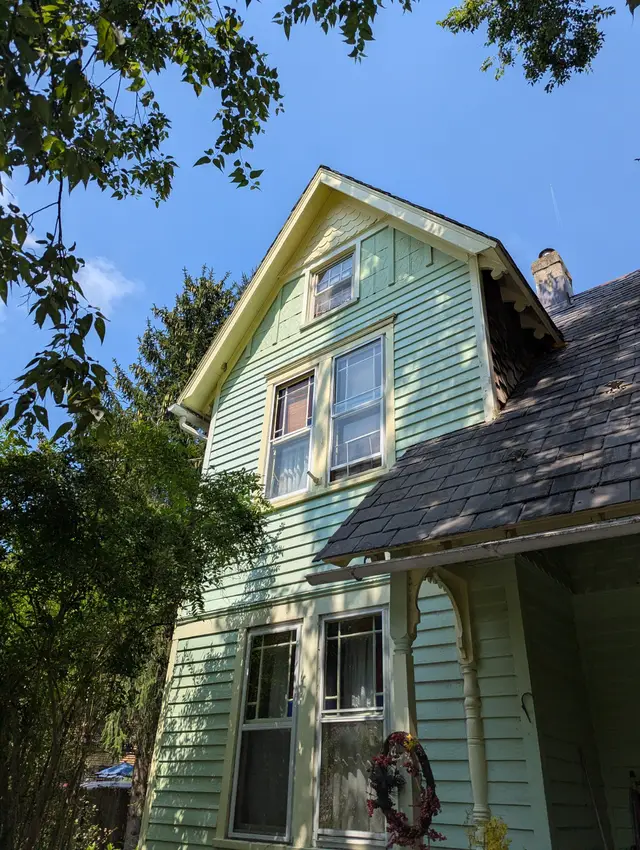 A pale green three-story Victorian with a steep slate roof, photographed from below through the trees.