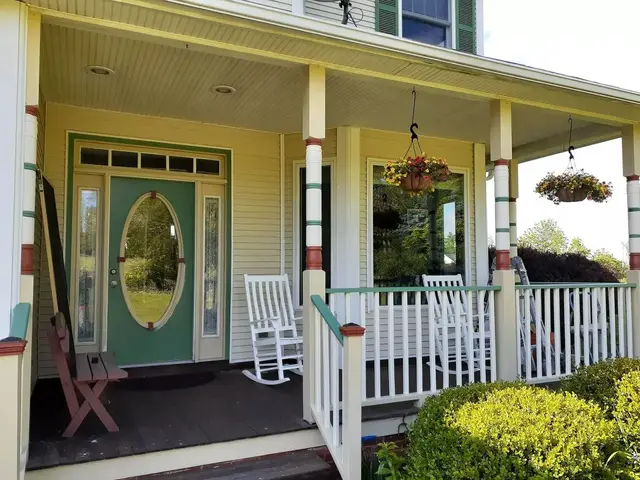 A Victorian front porch with white rails, painted posts, a green front door with an oval glass panel, and hanging baskets.