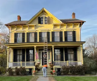 A yellow Italianate Victorian with black shutters and a deep porch; a painter works from a ladder at the porch.